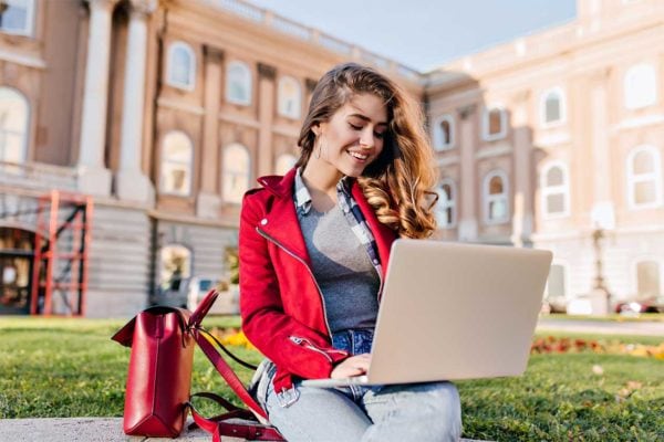Glamorous female student in red jacket sitting in the yard in front of college with computer. Charming freelance worker using laptop, chilling on the grass in warm day.. Glamorous female student in red jacket sitting in the yard in front of college with computer. Charming freelance worker using laptop, chilling on the grass in warm day.