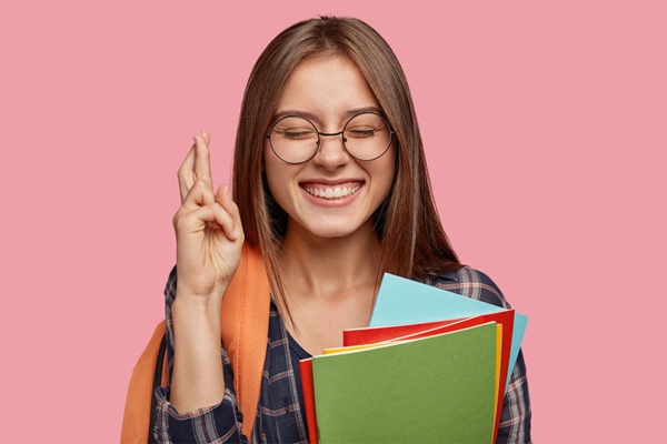 Intense cheerful schoolgirl with broad smile, crosses fingers for good luck, has satisfied expression, keeps eyes closed, carries rucksack, shows toothy smile, isolated over pink background. Intense cheerful schoolgirl with broad smile, crosses fingers for good luck, has satisfied expression, keeps eyes closed, carries rucksack, shows toothy smile, isolated over pink background.